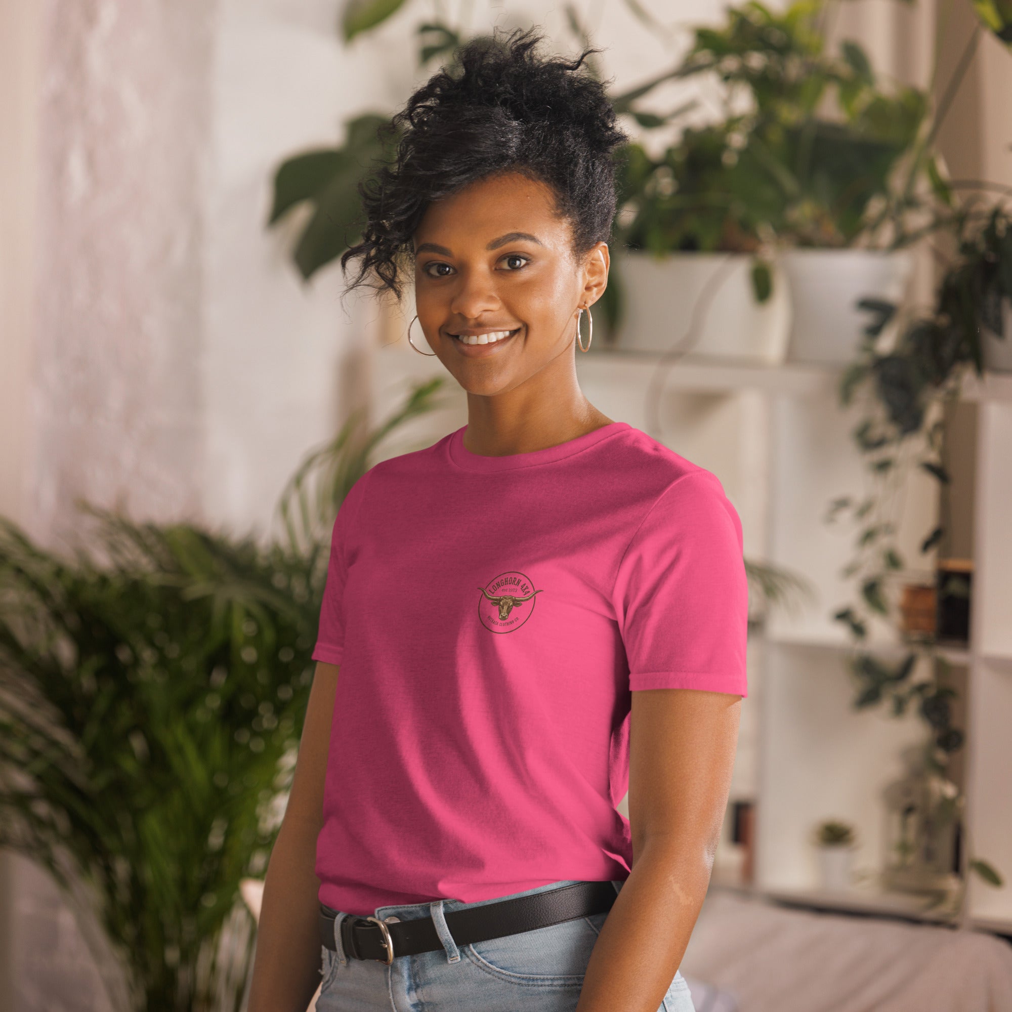 Person wearing a pink t-shirt with a logo, standing in a room with plants.
