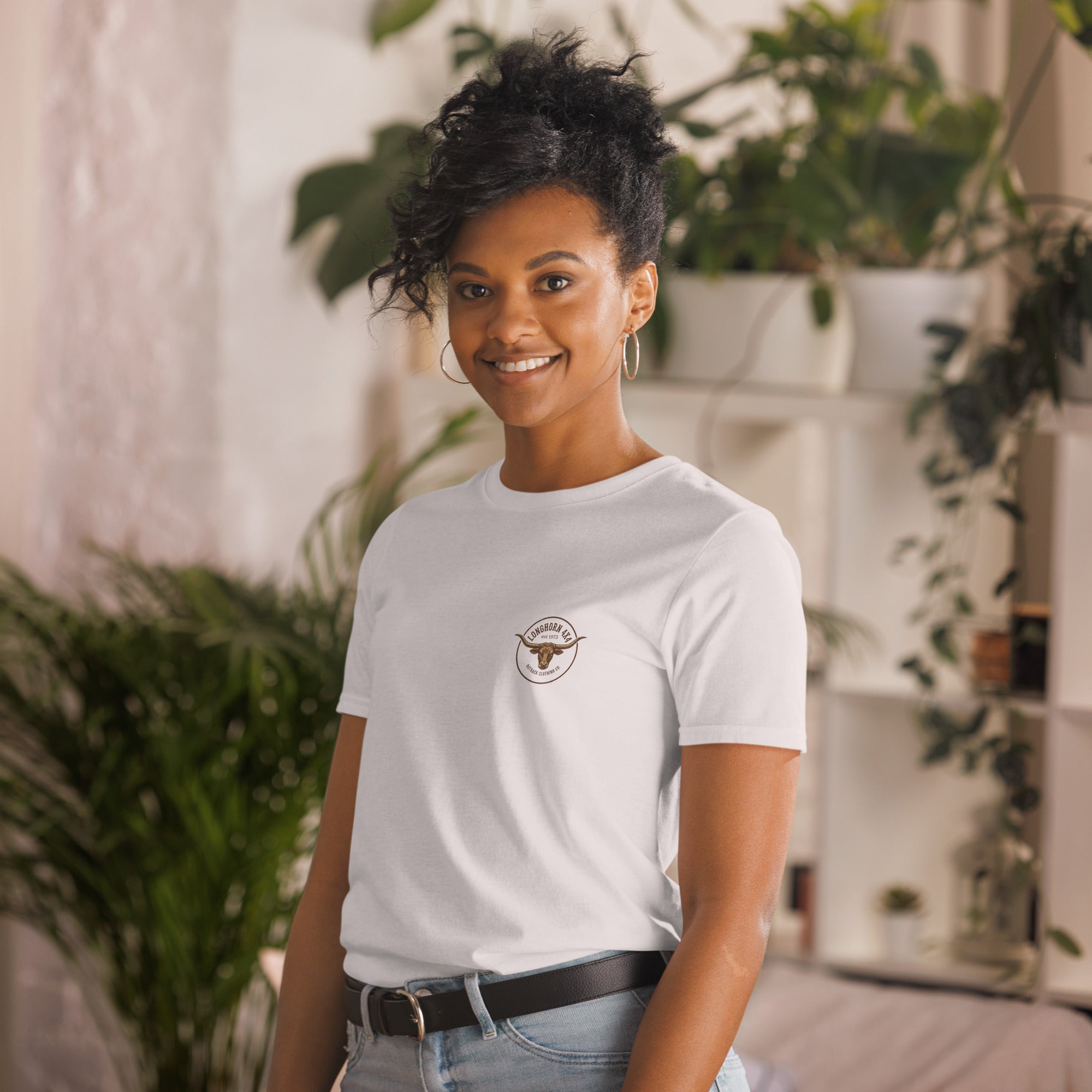 Woman wearing a white t-shirt with a logo, standing in a casual setting with plants.