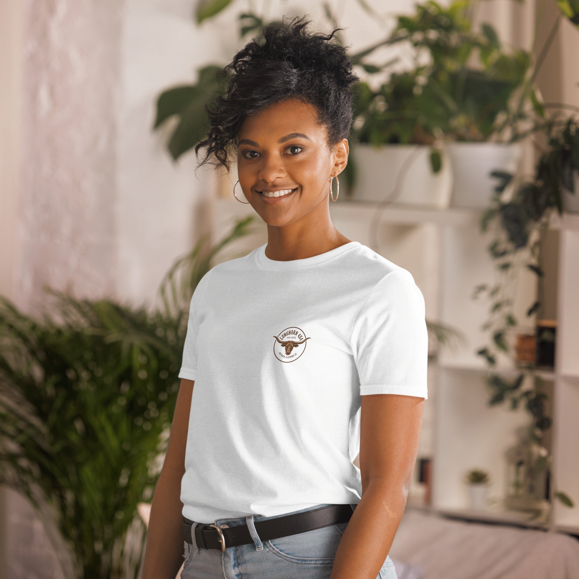 Woman wearing a white t-shirt with a logo, standing in a casual setting with plants.